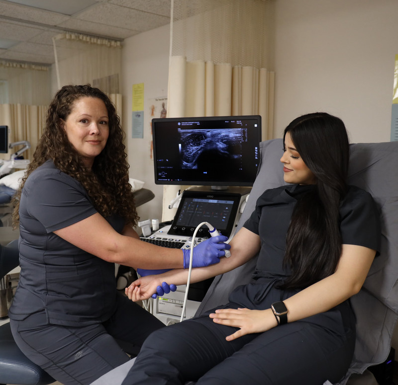 A student in training performs a sonography over their patient's inner elbow.